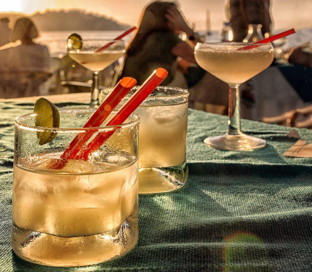 beach bar scene, with margarita cocktail drinks on a table, and people on the background watching the sunset on the ocean