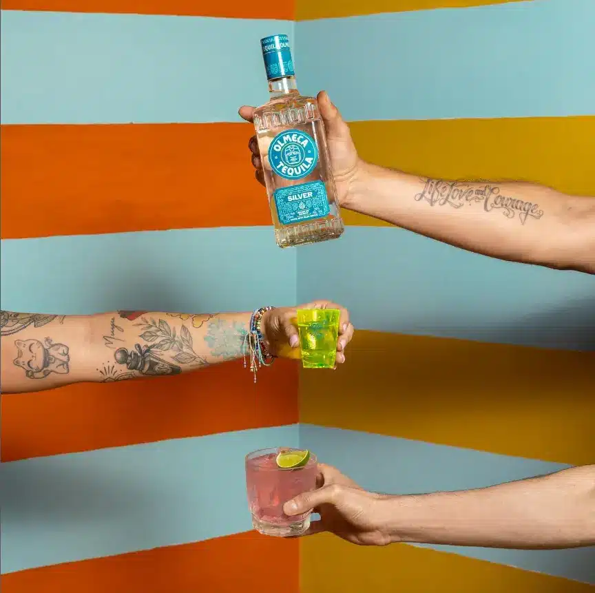 Friends at a sneakers party hold Olmeca Tequila bottle a cocktail and a shot of Olmeca Tequila at a party